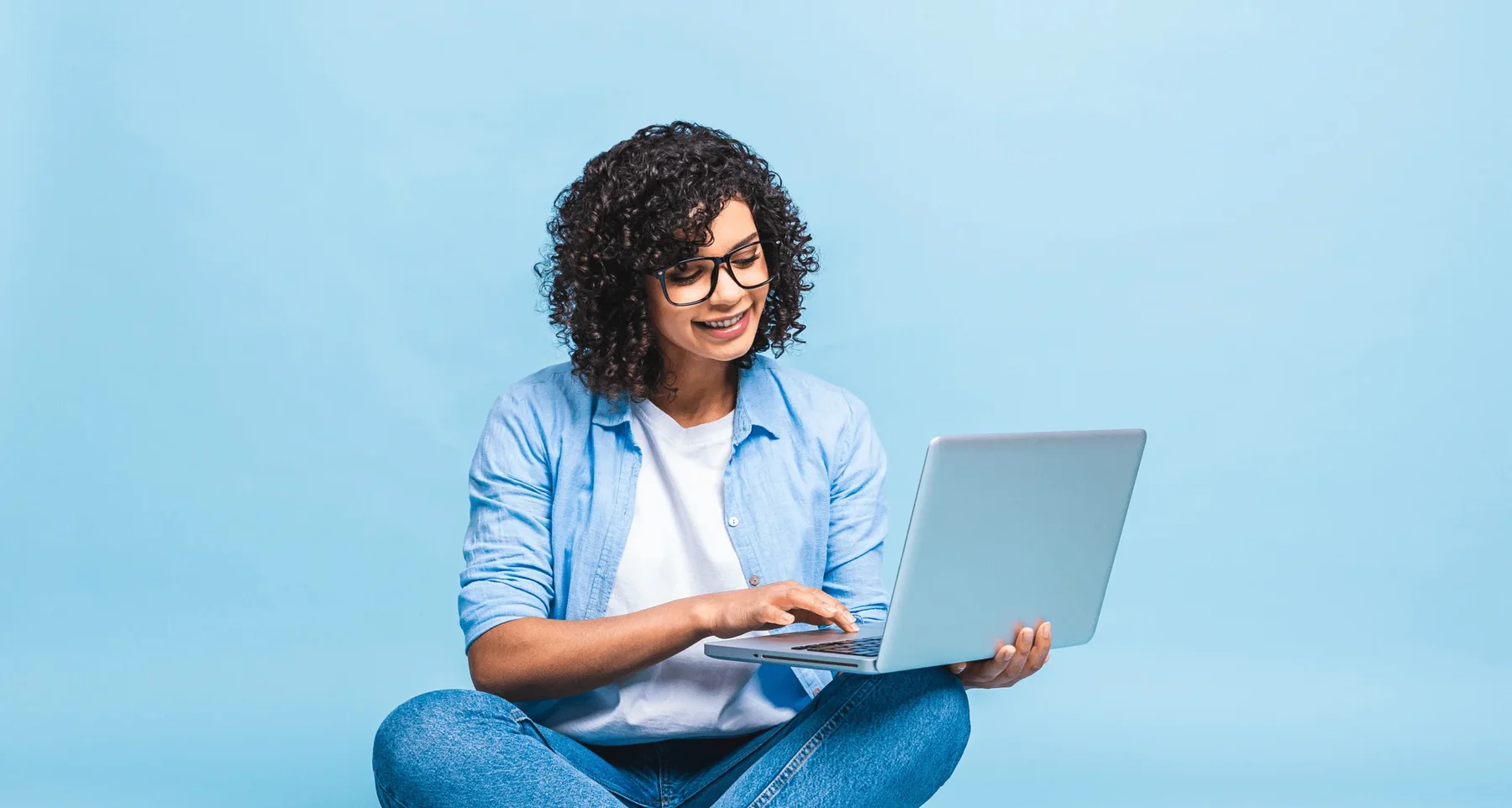 portrait-african-american-black-woman-casual-sitting-floor-lotus-pose-holding-laptop-isolated-blue-background-scaled